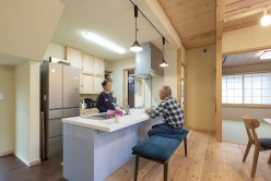 木製のカウンターキッチンで会話を楽しむ高齢の夫婦。和室が奥に見える。